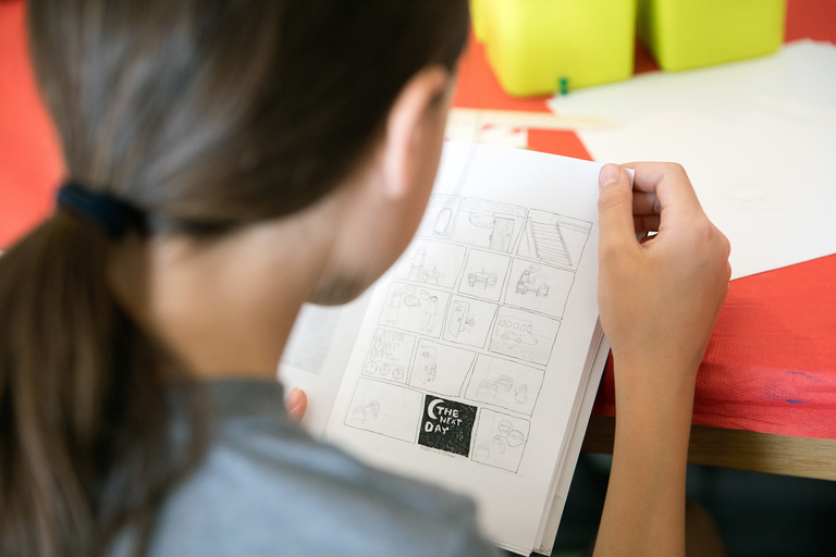 An over-the-shoulder view of a young girl with her student-designed comic strip, created during the IYWP Summer Camp